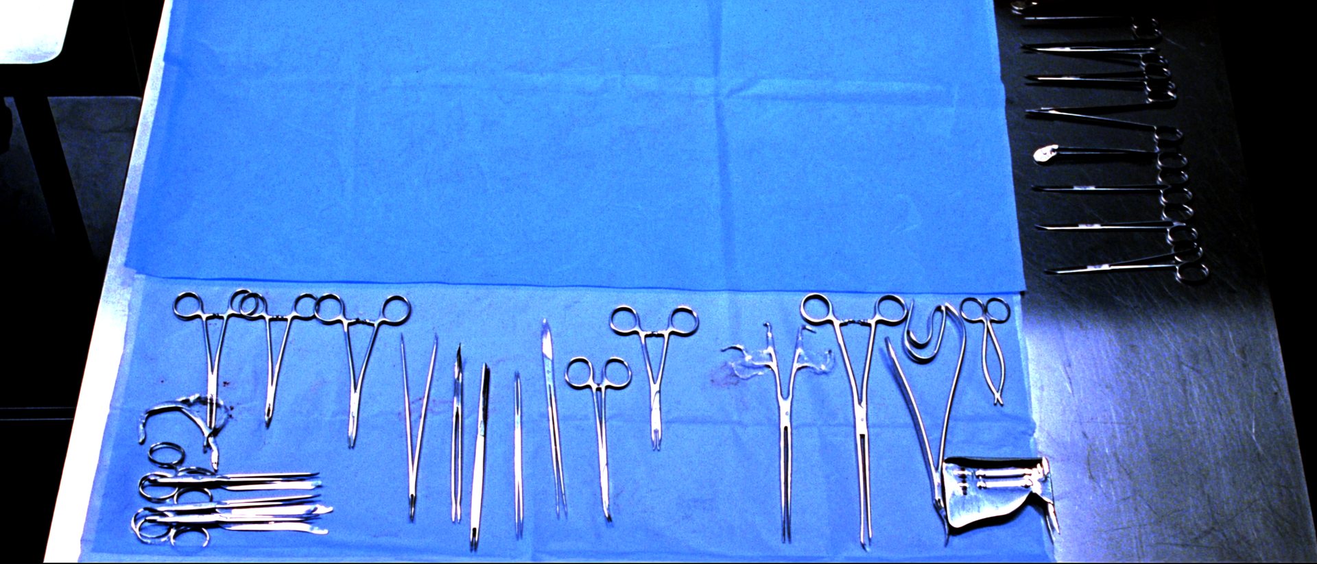 Stainless steel surgical instruments laid out in precise parallel rows on a folded blue sterile drape on a steel table, lit evenly from overhead.