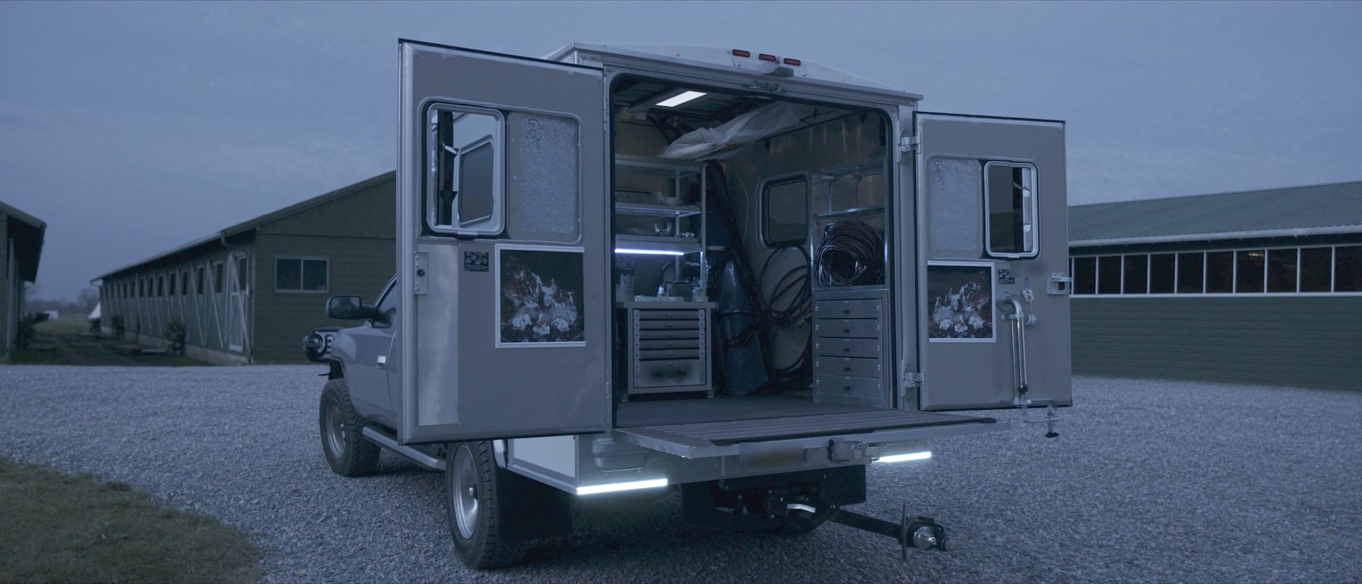 The open rear of a box-body veterinary field unit, both rear doors swung wide, the shelved and drawered interior lit by a strip light, parked on gravel between two long low barns at dusk.